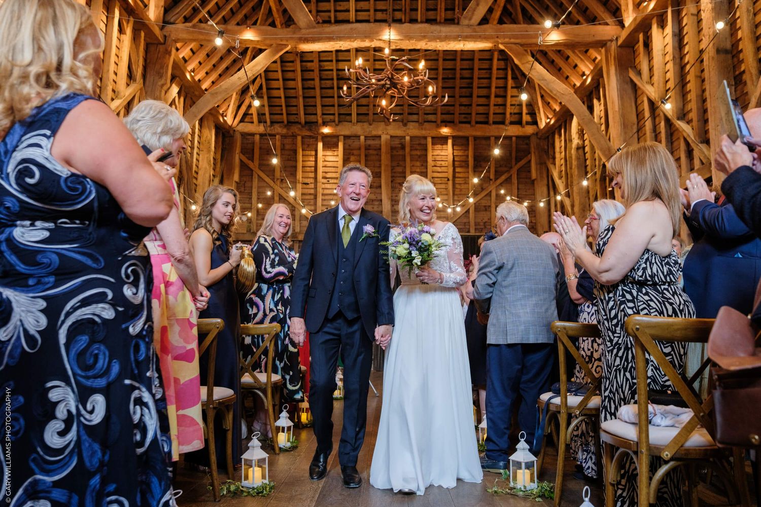 Wedding ceremony taking place in a rustic wooden barn venue with exposed beams and hanging chandeliers.