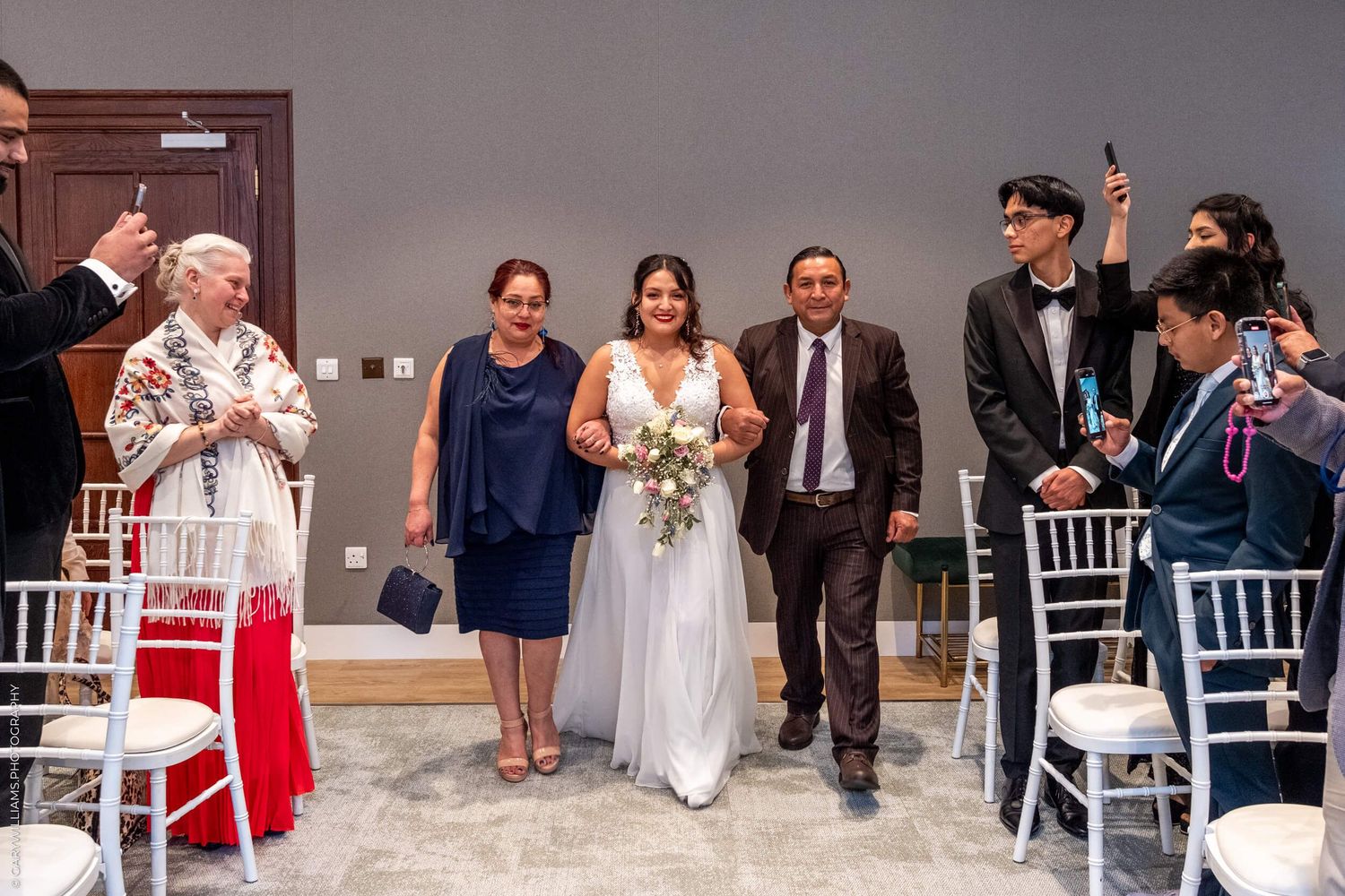 Wedding ceremony processional in a modern indoor venue with white chiavari chairs lining the aisle.
