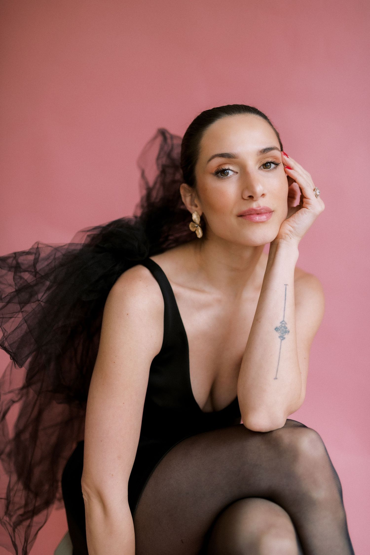 Portrait against a pink backdrop featuring a dreamy tulle dress, dark tights, and delicate earrings in a soft studio setting.