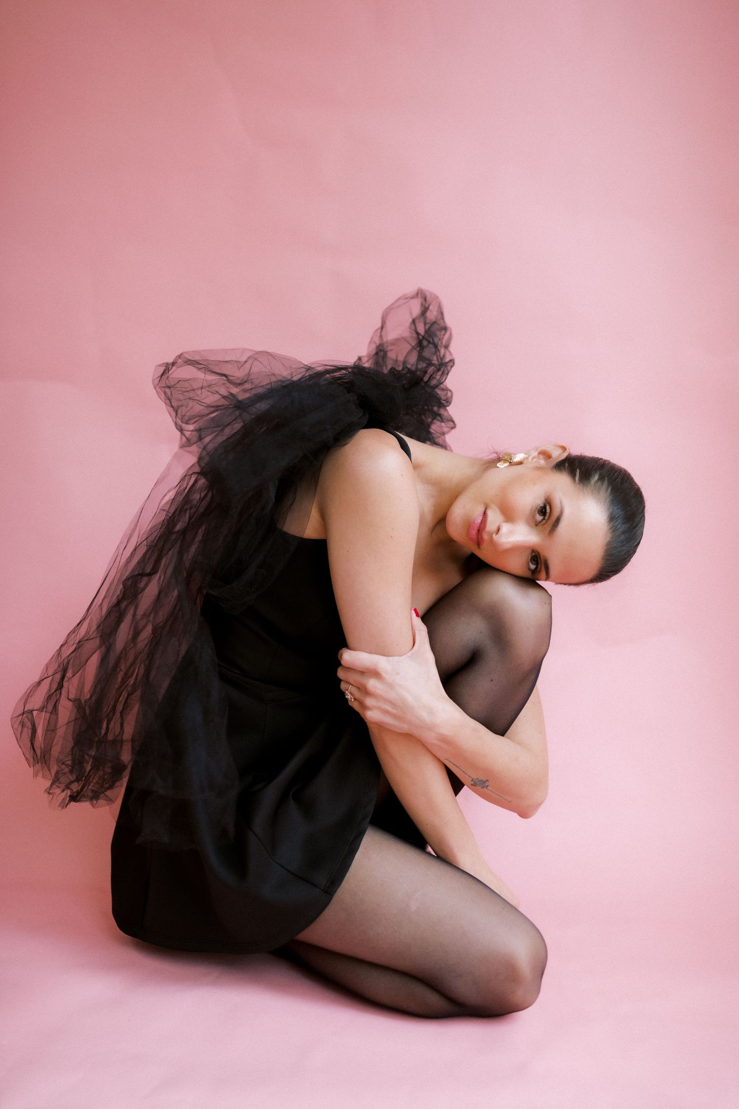 A dancer in a black tulle dress and tights poses gracefully against a pink backdrop during a studio photoshoot.