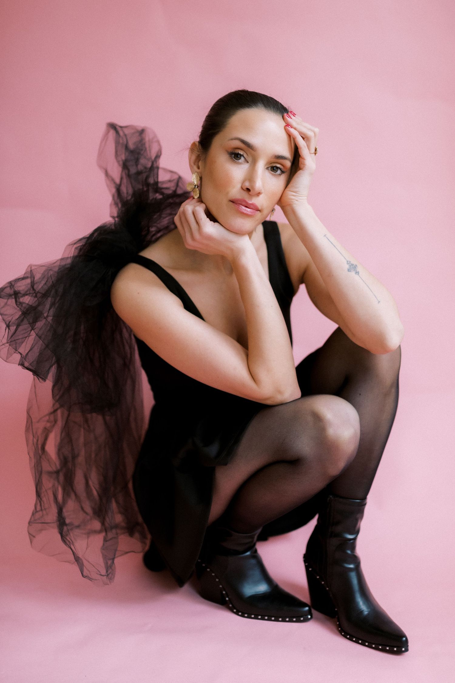 Figure in black tulle dress and boots poses gracefully against pink backdrop in artistic portrait.