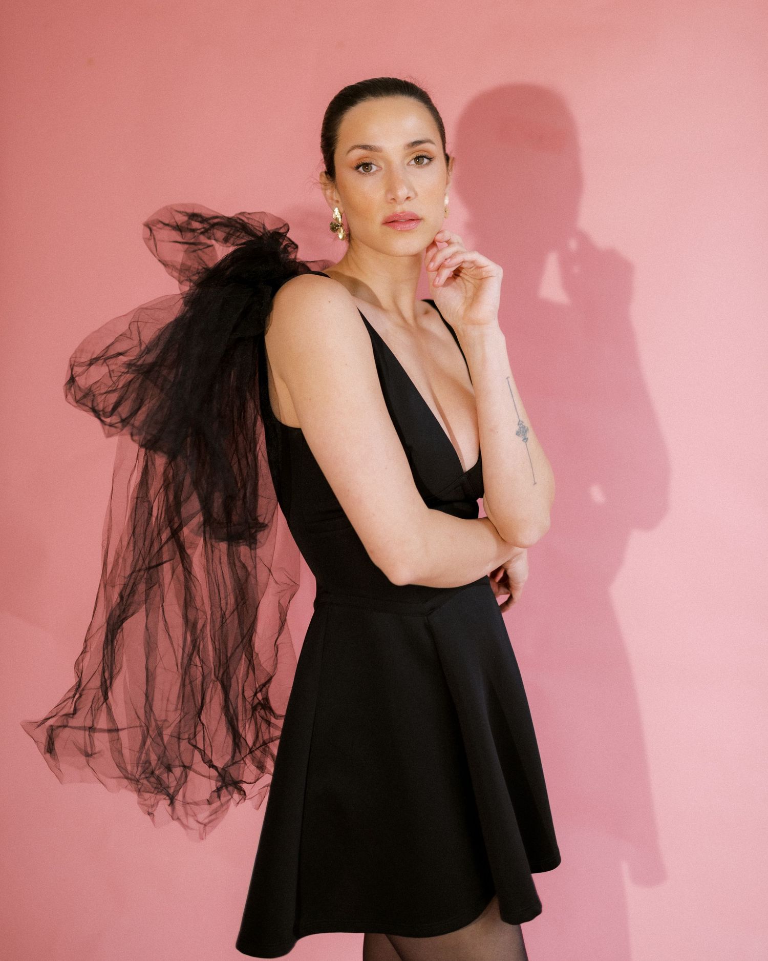 Fashion model in flowy black dress poses against pink wall with dramatic tulle fabric flowing behind.