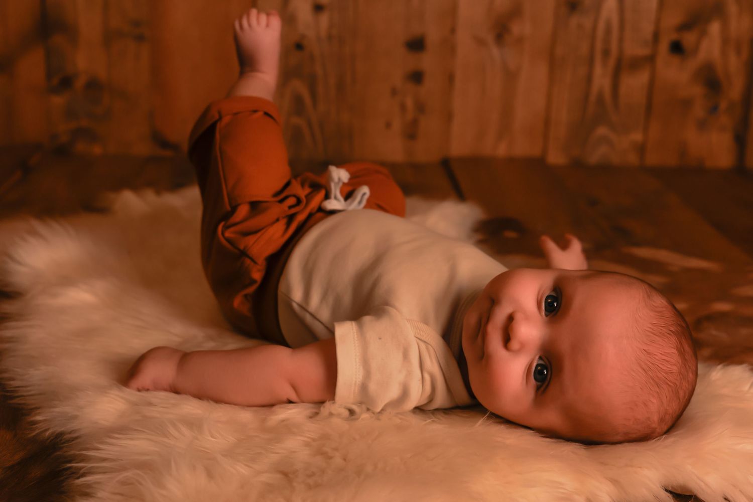 A baby in brown and white clothing lies on soft white fur against wooden paneling.