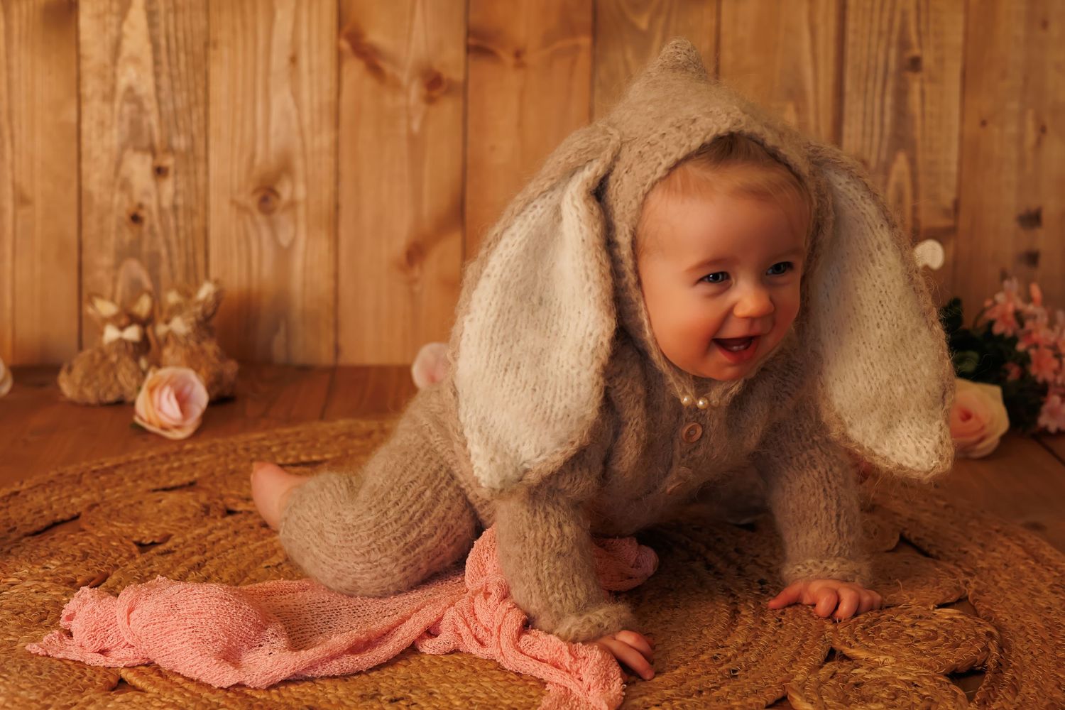 A small child wearing a fuzzy grey bunny costume crawls on carpet near pink fabric and wooden walls.