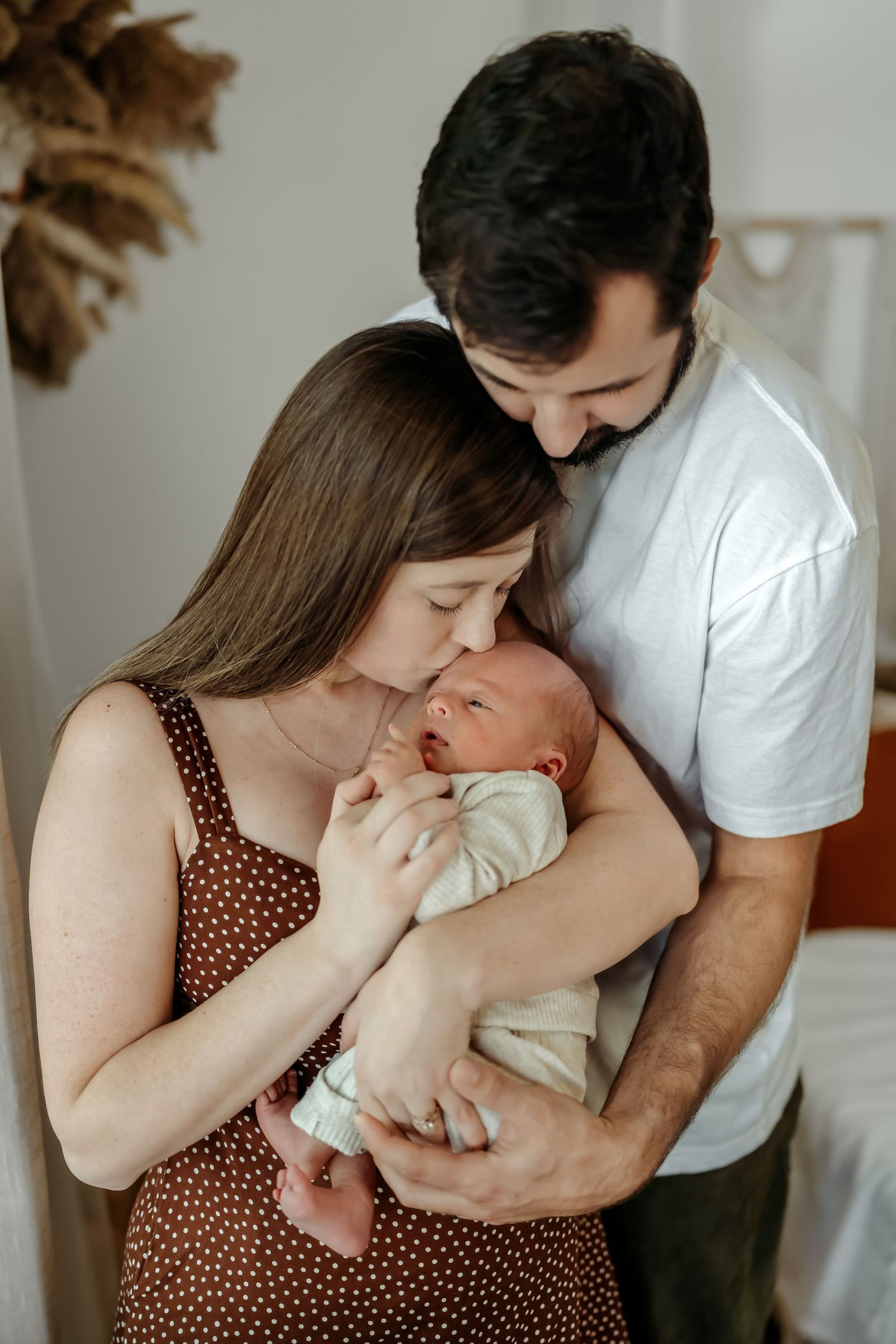 Parents in white and polka dot dress hold and gaze lovingly at their newborn baby in a tender family moment.