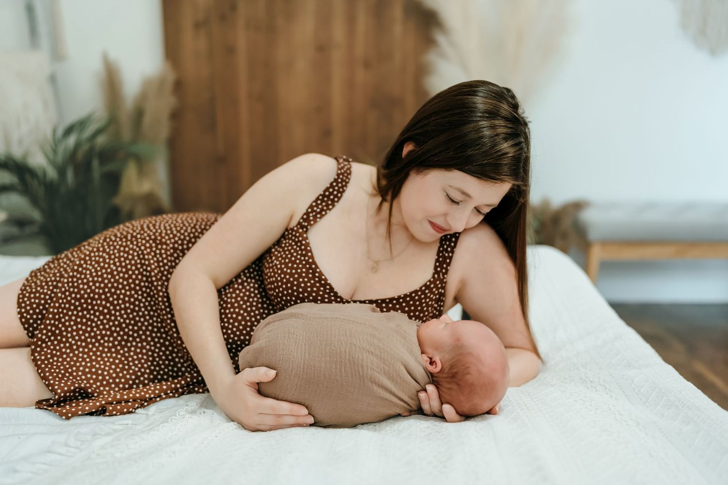 A mother in polka dots nurses her baby while lying down on a white bed in a softly lit room.
