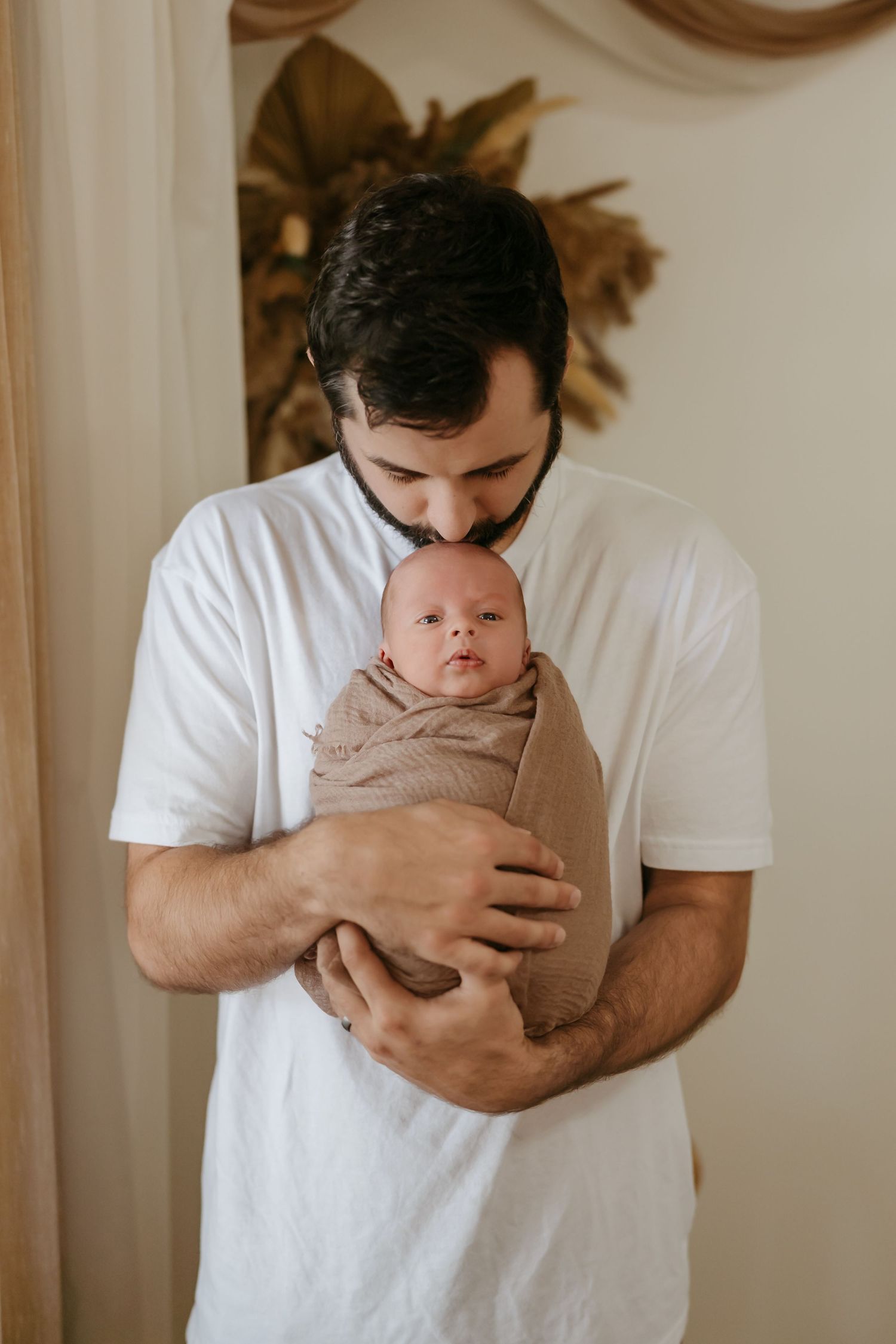 Parent in white t-shirt tenderly holds swaddled newborn baby wrapped in beige fabric against decorated wall.