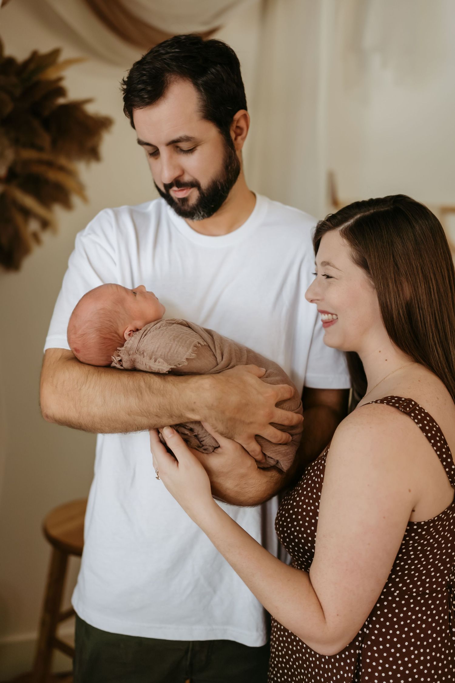 Parents lovingly hold their newborn baby in a candid indoor photo with warm lighting and pampas grass decor.