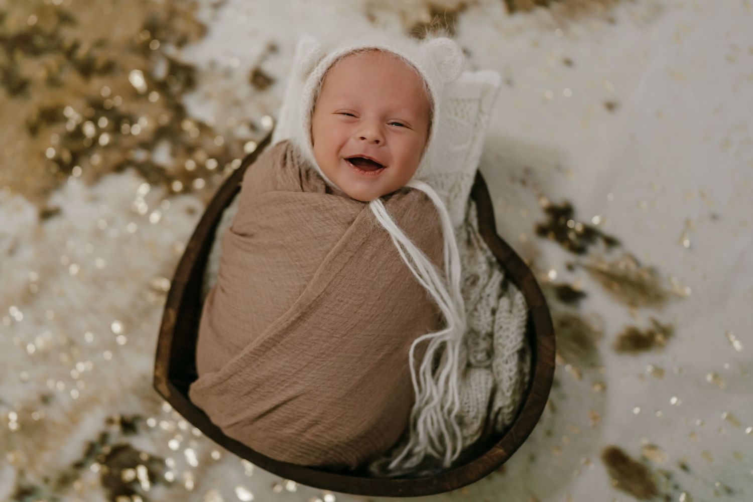 Smiling baby wrapped in brown swaddle lying in dark basket against sparkly background.