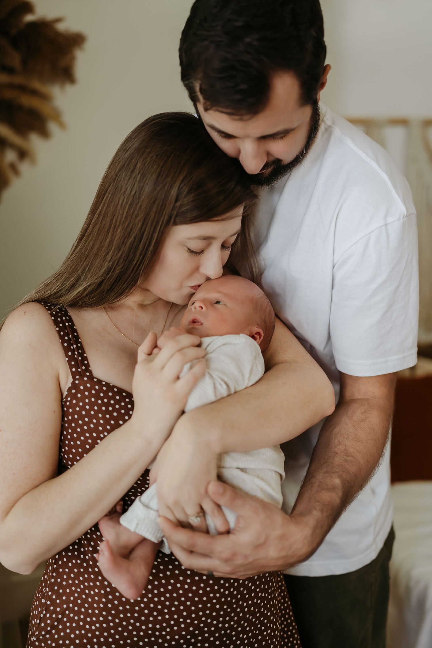 Parents tenderly hold and kiss their newborn baby while standing together in a cozy room.