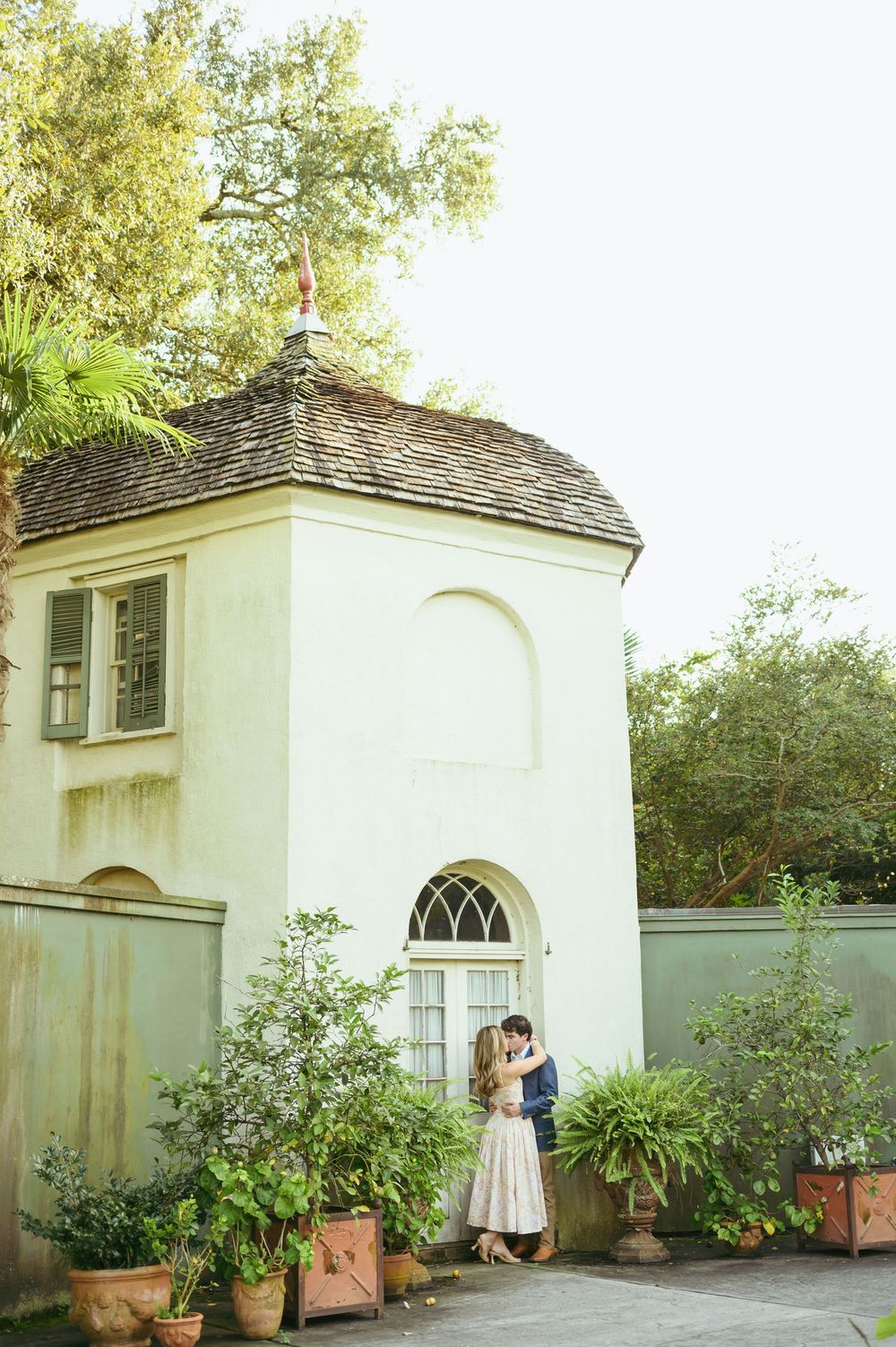 A charming white garden house with an arched doorway surrounded by potted plants and greenery.