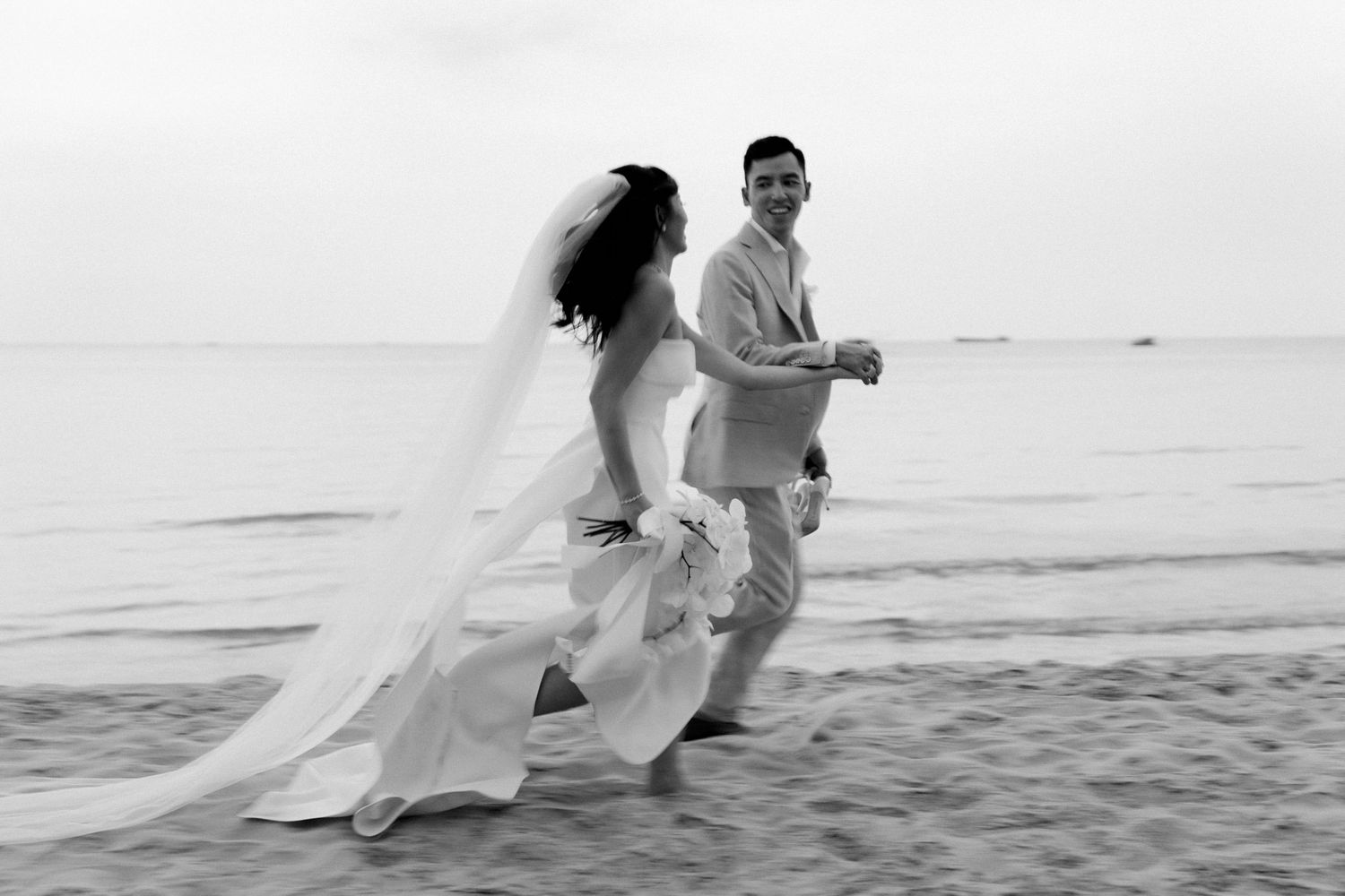 A romantic sequence of a couple running playfully together on the beach in elegant white attire at sunset.