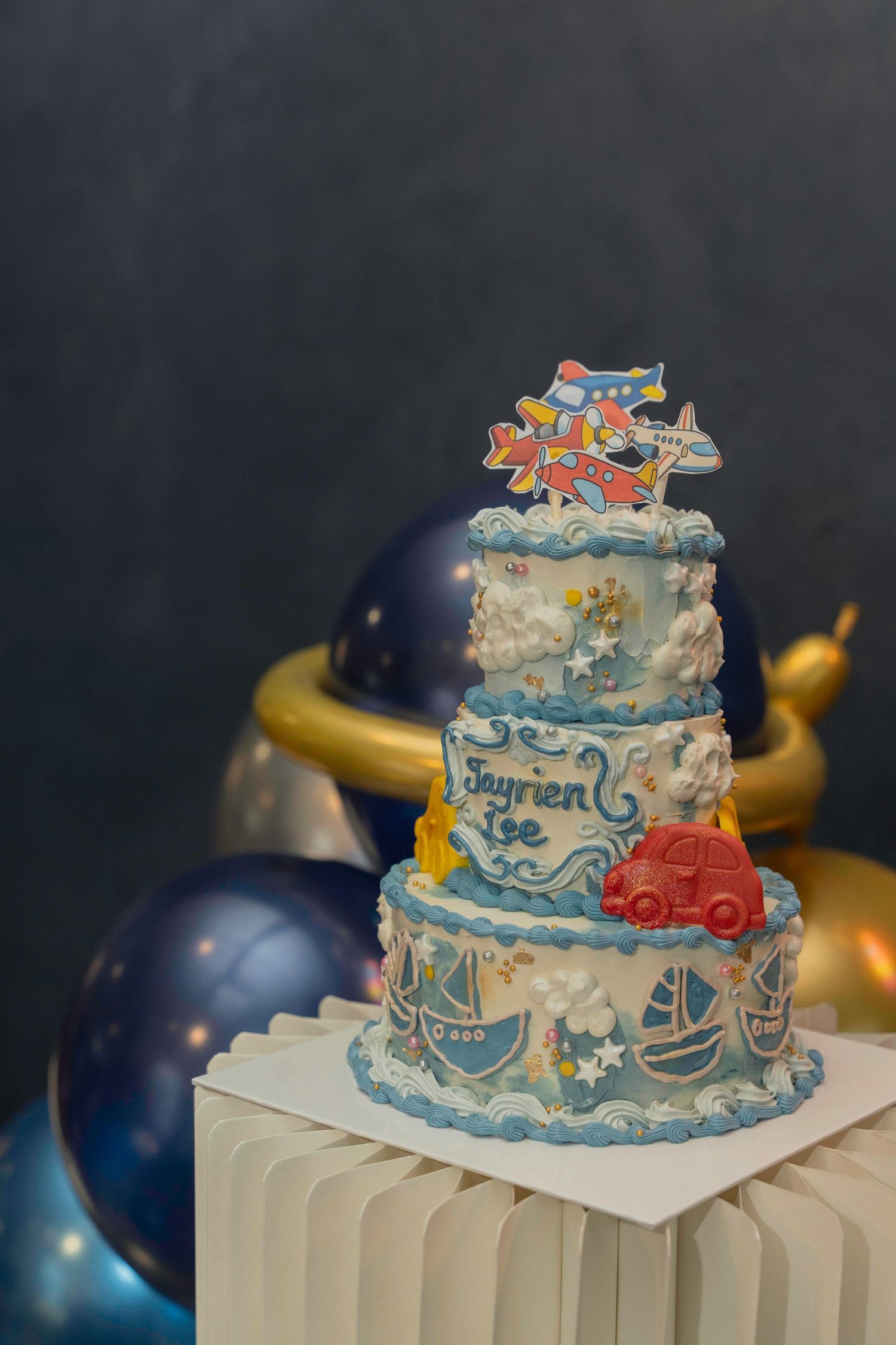A blue and white birthday cake decorated with maritime-themed designs sits atop white pleated paper with dark balloons nearby.