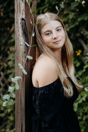 Portrait photos in a black off-shoulder top against green foliage and wooden post background.