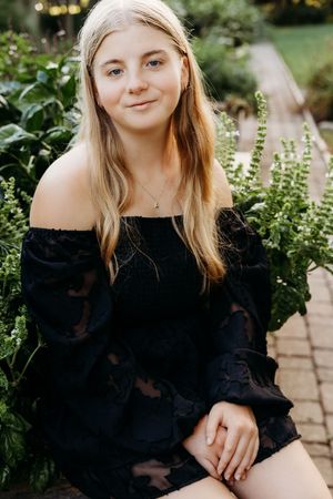 Portrait in a garden wearing a black off-shoulder dress with long sleeves amidst greenery and flowers.