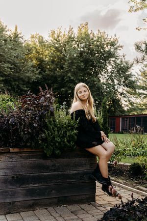 Portrait in a garden setting with rustic wooden planters, wearing a black dress and boots at sunset.