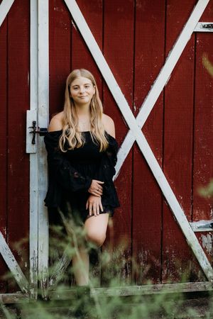 A scenic portrait against a red barn door with white trim, wearing a black dress in natural setting.
