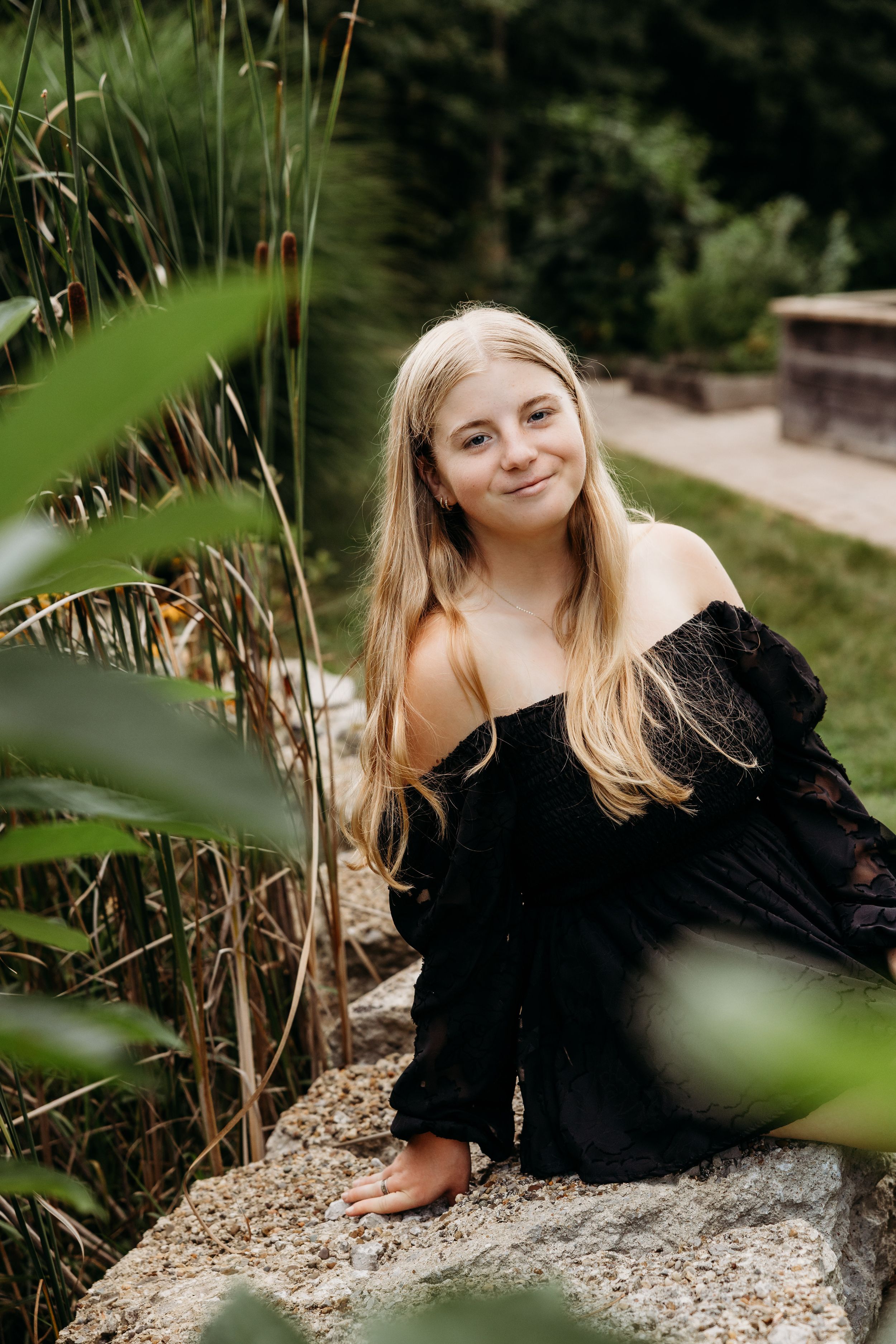Portrait in a garden setting with black off-shoulder dress amid lush greenery and stone features.