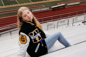 Someone poses casually in a black varsity jacket and jeans while sitting on red bleachers at a sports venue.