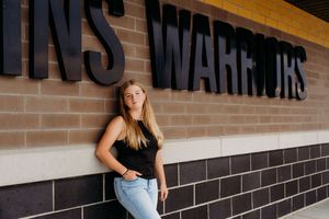 Woman in black tank top and jeans leans against brick wall with typography signage above.