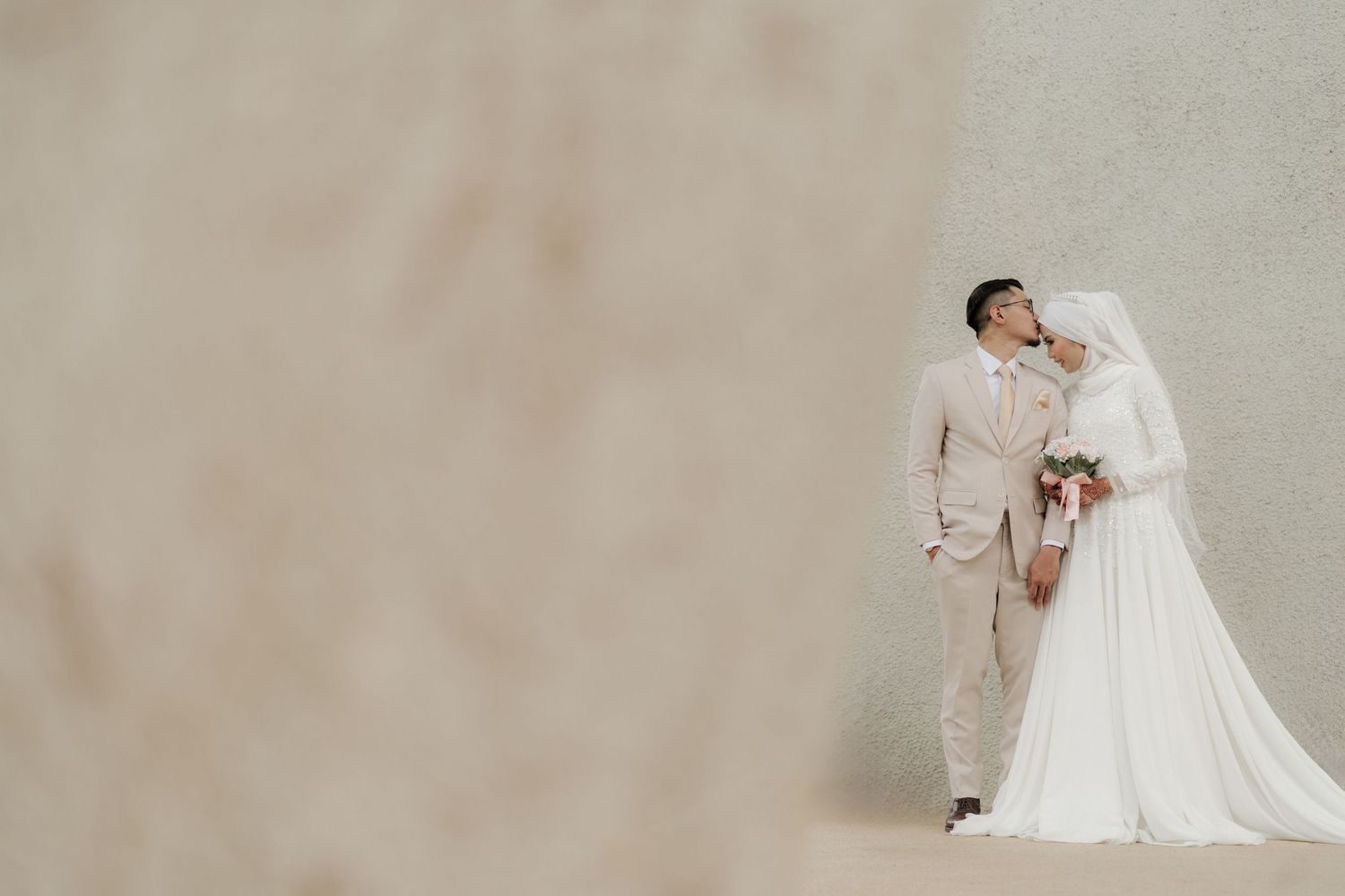Elegant couple in beige suit and white wedding gown pose against a minimalist backdrop.
