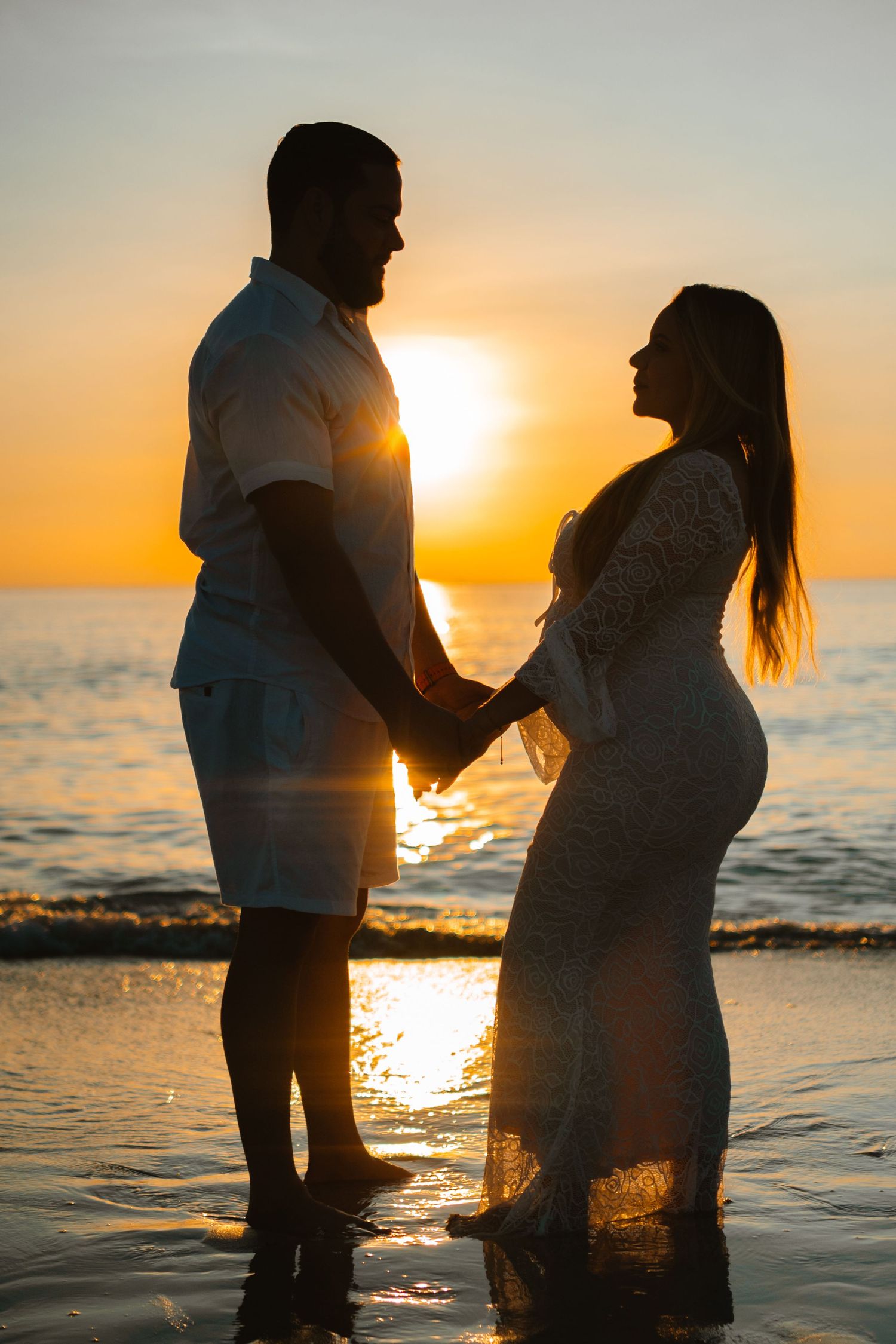 Silhouetted couple stands together at beach during golden sunset with water reflecting orange sunlight.