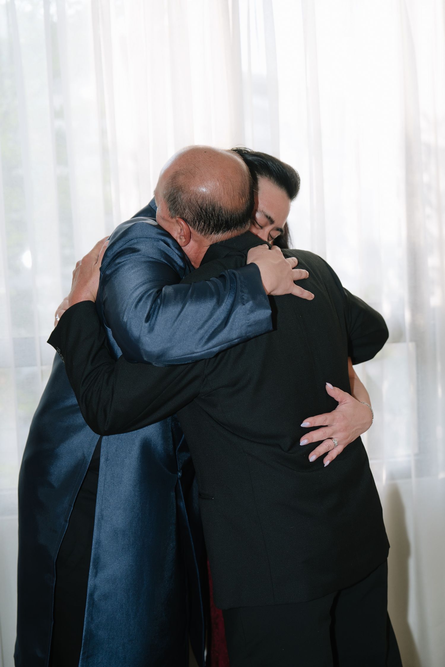 Two men in formal wedding attire sharing an emotional embrace by sheer curtains.
