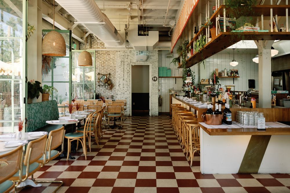 Bright bistro interior with checkered floors, rattan bar stools, and lush greenery decor.