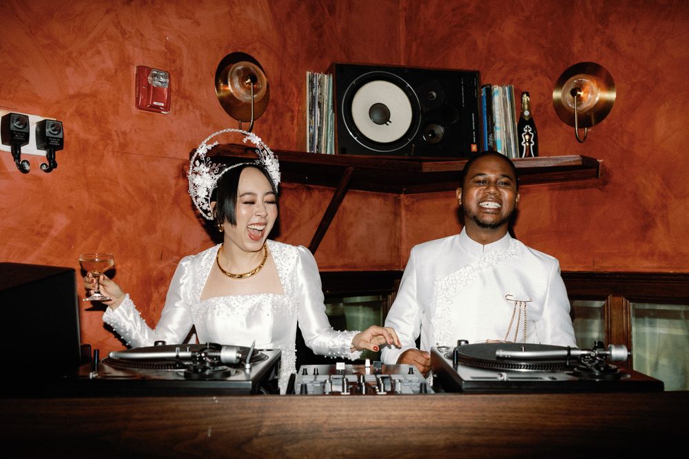 Bride and groom DJing together at turntables in a warmly lit red-walled venue.