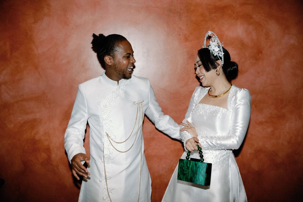Newlyweds in matching white outfits laughing together against a warm terracotta-colored wall.