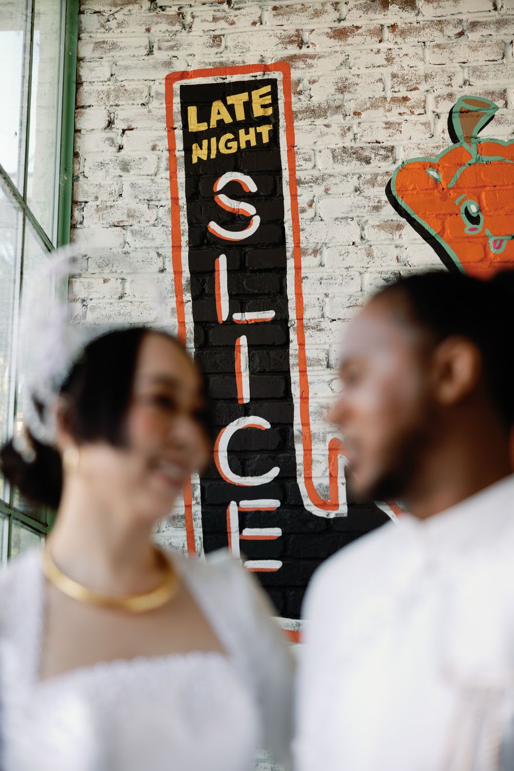 Bride and groom blurred in foreground before a Late Night Slice painted brick sign.