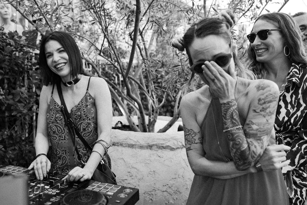 Three women laughing joyfully outdoors at a wedding reception, one operating a DJ setup.