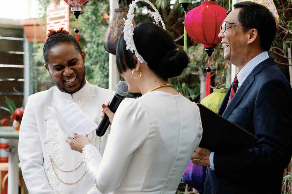 Groom laughing joyfully during outdoor wedding ceremony while bride speaks into microphone.