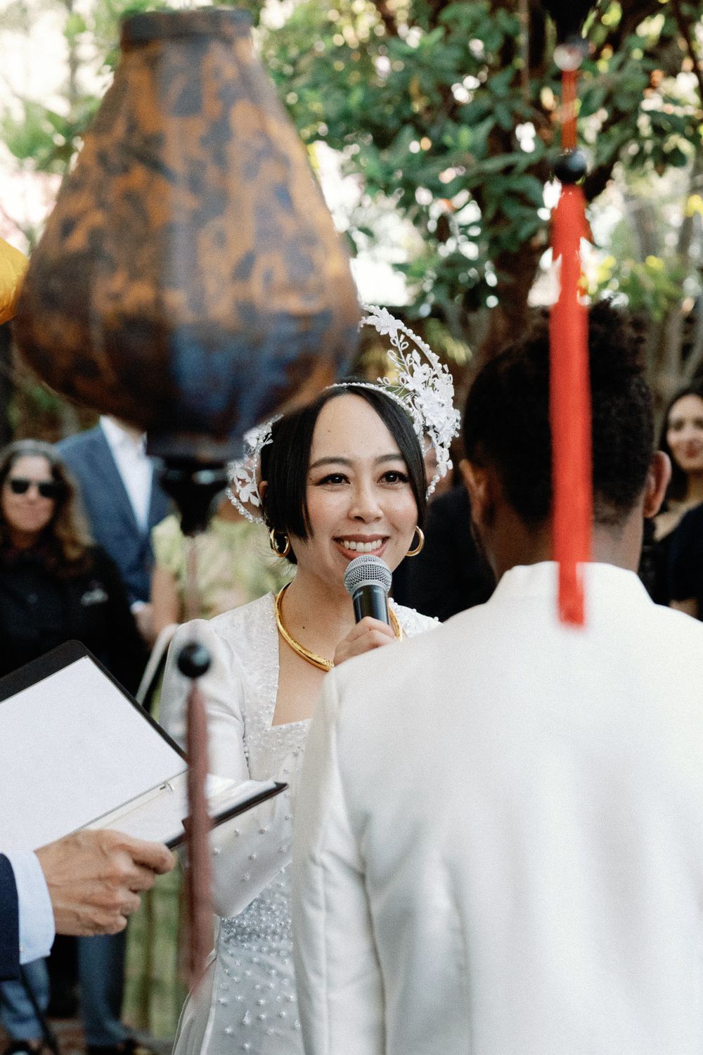 Asian bride in white ao dai and floral headpiece smiling while reciting vows at outdoor ceremony.