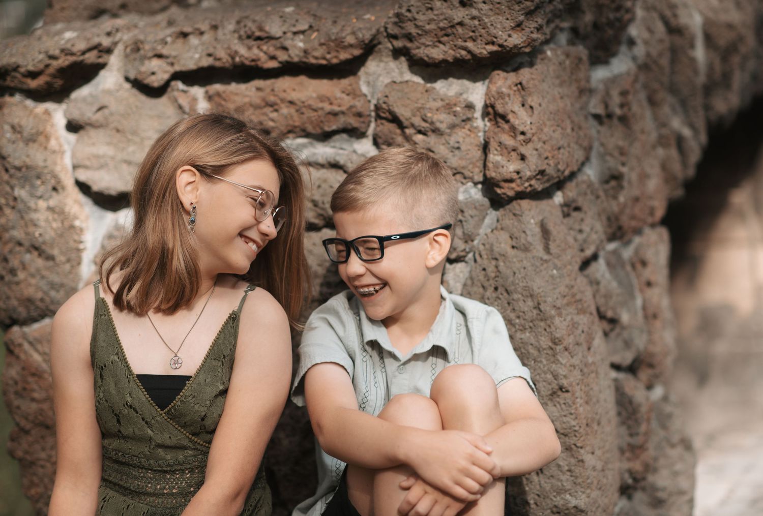 Siblings leaning against a stone wall at Manito Park, laughing at each other being silly during a family photo session in Spokane.