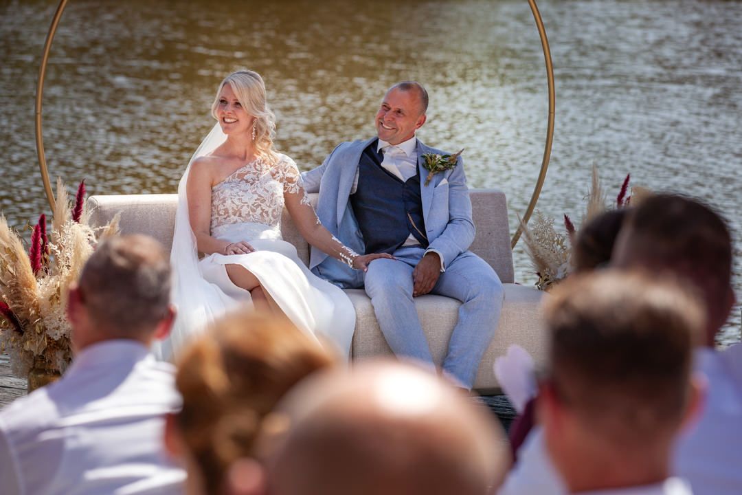 Overzicht van de trouwceremonie bij L Seven met het bruidspaar aan het water en gasten op de voorgrond