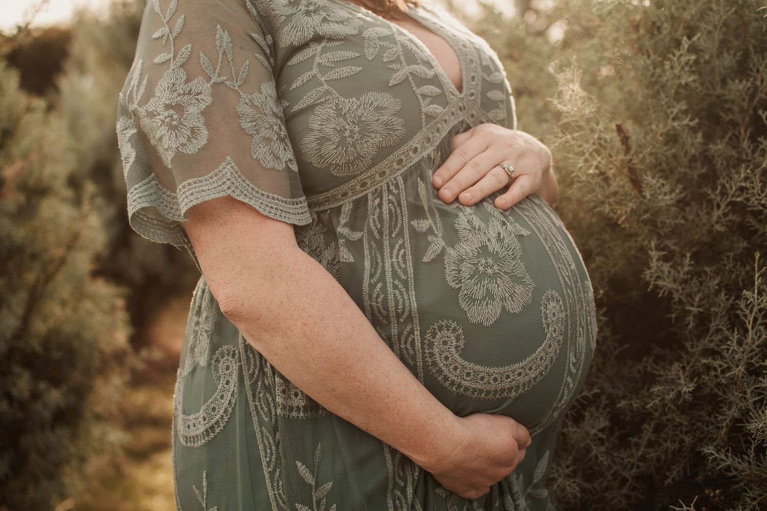 A pregnant woman in a green lace dress holds her baby bump during a golden hour maternity photoshoot outdoors.