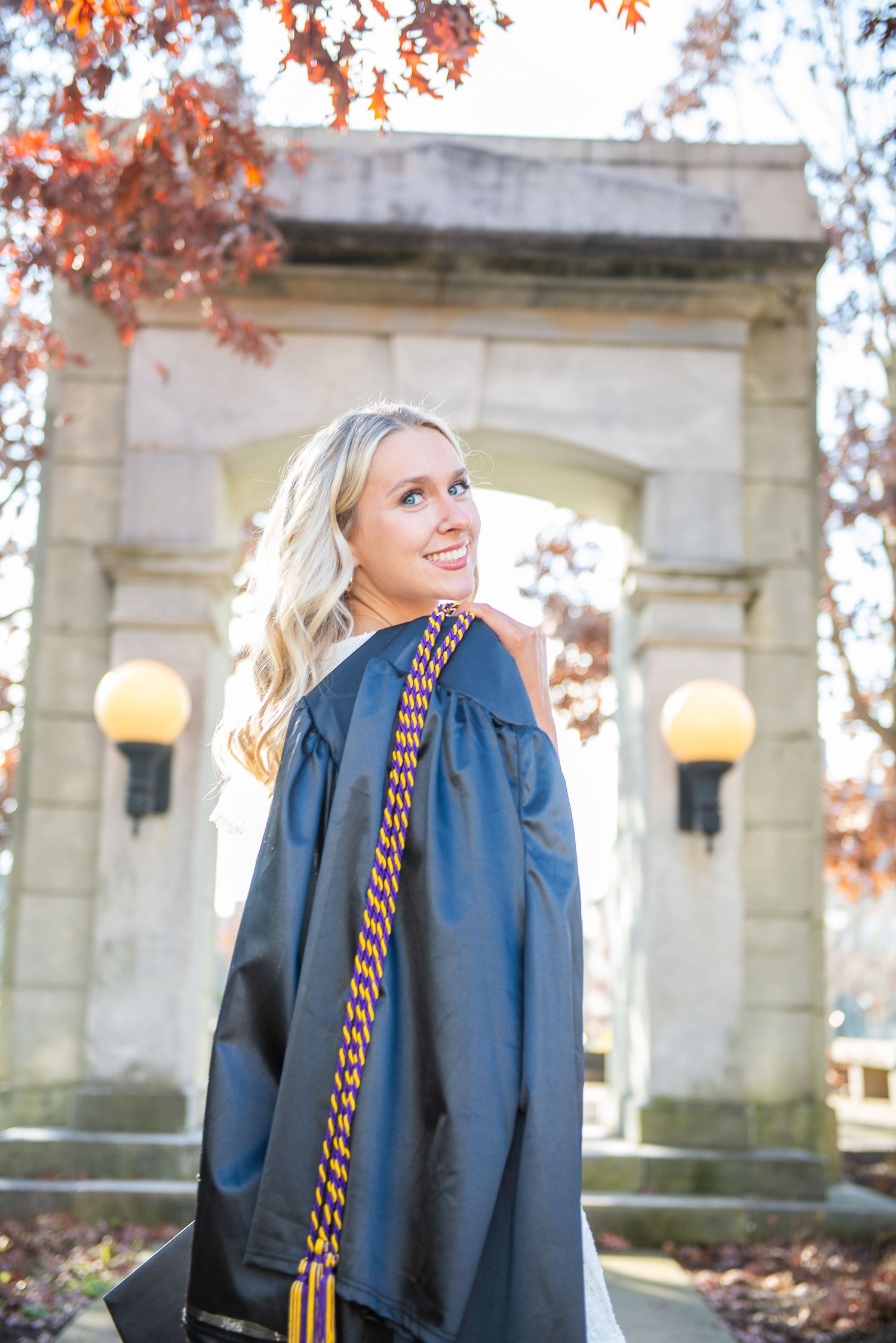 Graduate in cap and gown with purple and gold honor cords poses outdoors on campus during autumn.