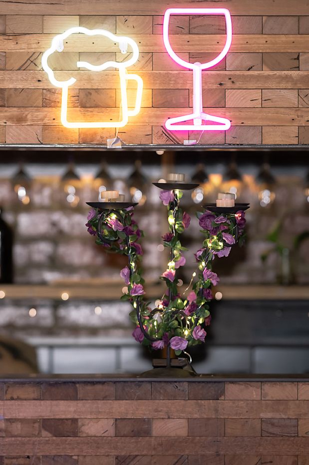 Neon wine glass and beer signs illuminate a rustic wooden wall with decorative floral vines and string lights.