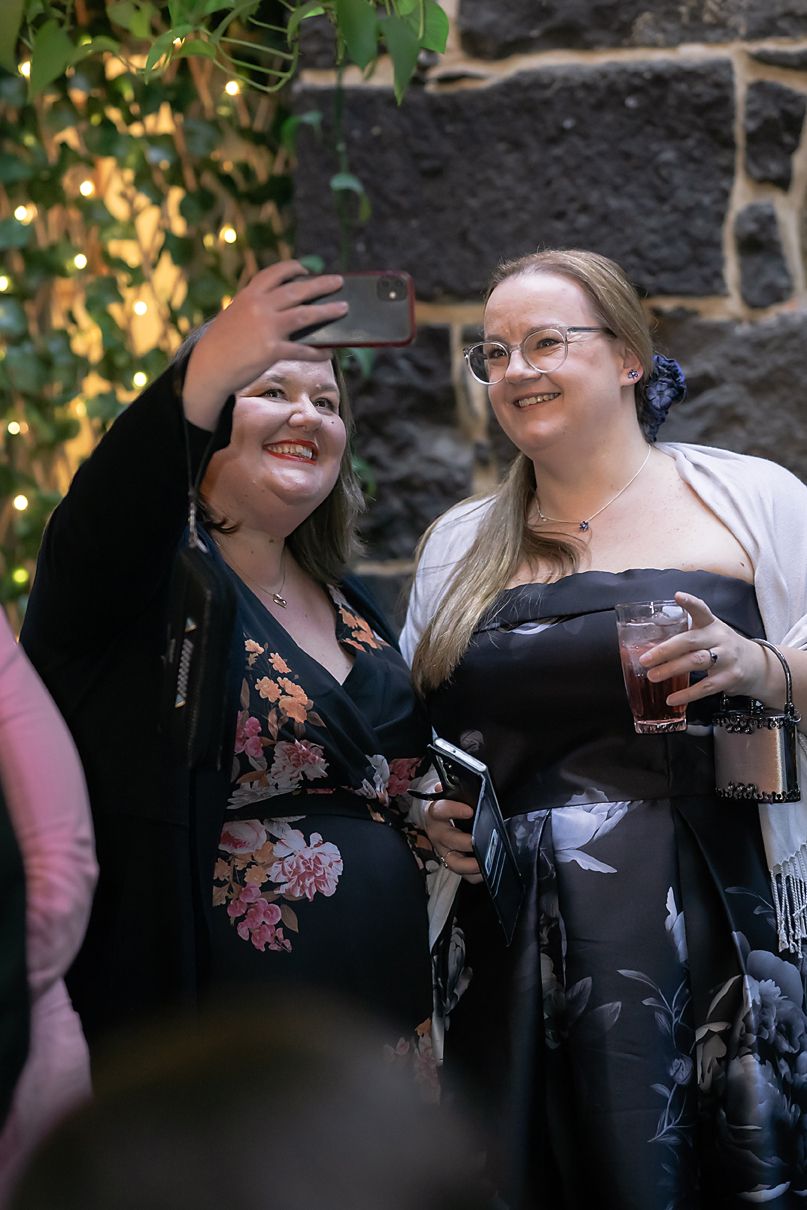 Candid sequence of two friends taking selfies together while laughing outdoors in black outfits.