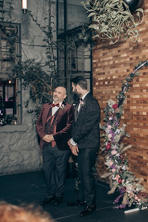 Two people in formal attire share a candid moment against a brick wall adorned with floral decorations.