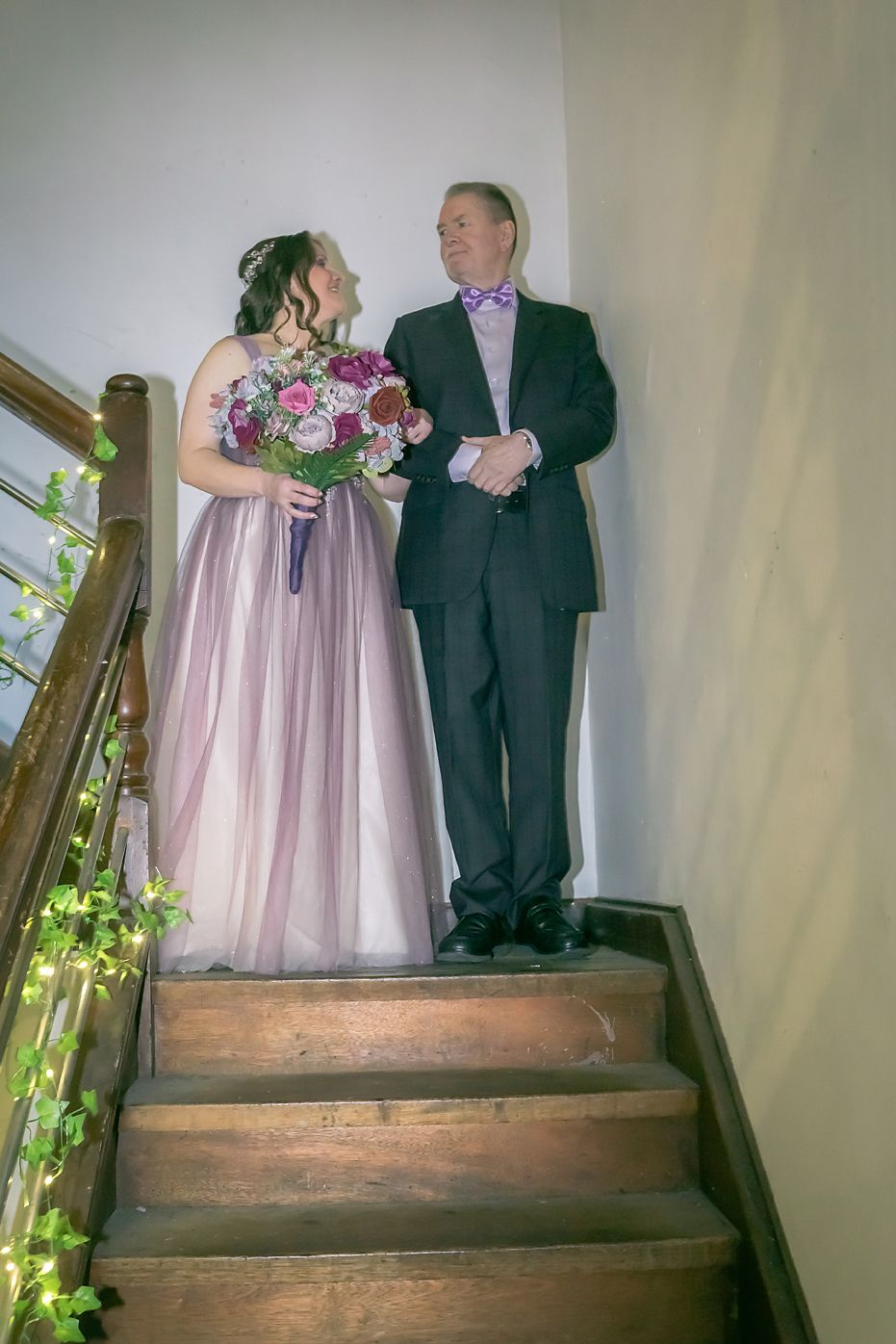A couple in formal attire stands on rustic wooden stairs with decorative greenery on the banister.