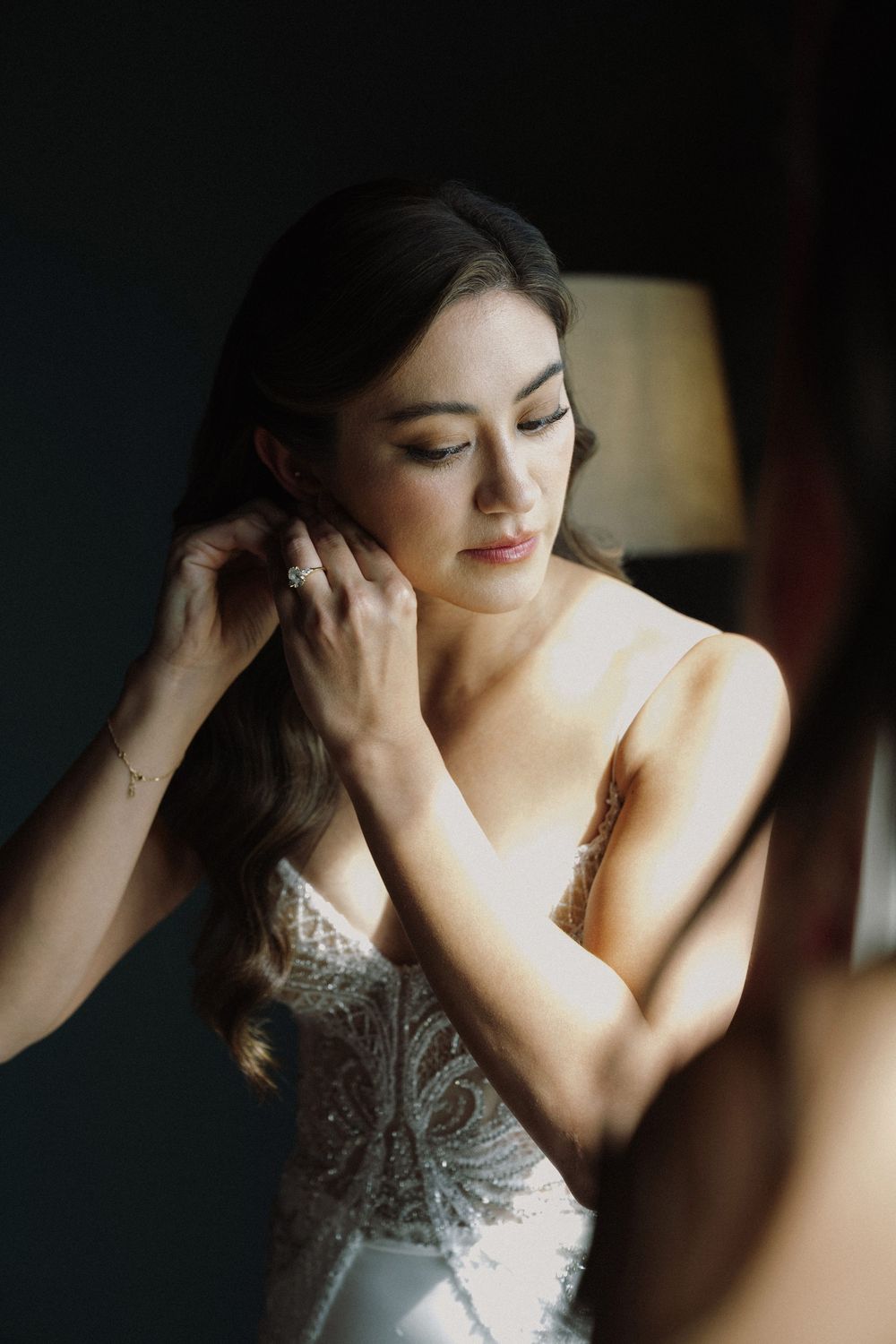 A woman in an elegant beaded white wedding dress adjusts her hair in soft natural lighting.