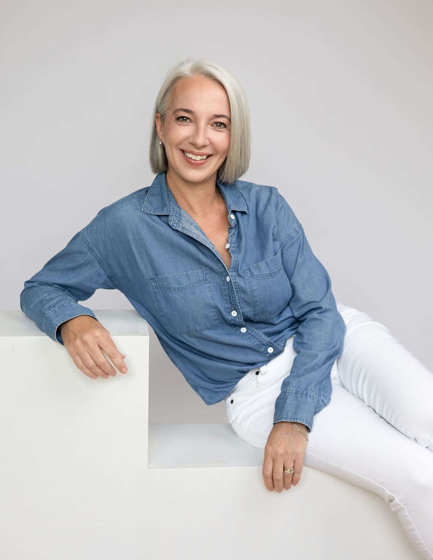 Short silver haired woman with big smile wearing blue button down shirt with white posing in a leaned sitting pose on a white backdrop.