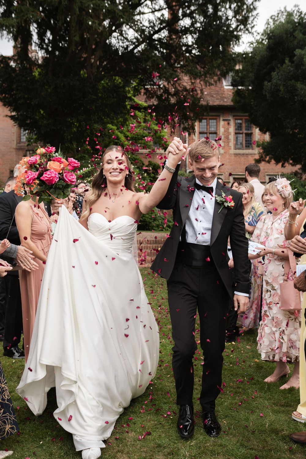 Joyful wedding couple celebrates while walking through shower of rose petals on garden lawn with guests looking on.