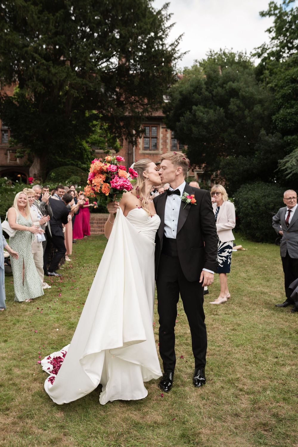 Wedding party celebrates outdoors on a grassy lawn with guests throwing flower petals in celebration.