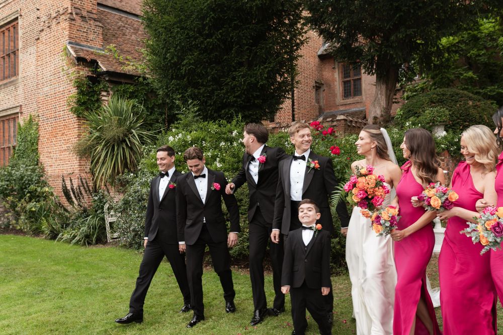 Wedding party members in formal attire walk and laugh together on the lawn of a brick building.