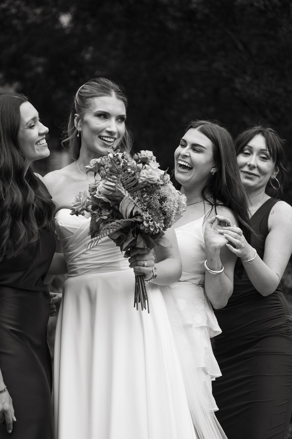 A wedding party in black and white dresses share joyful laughter while holding bouquets.