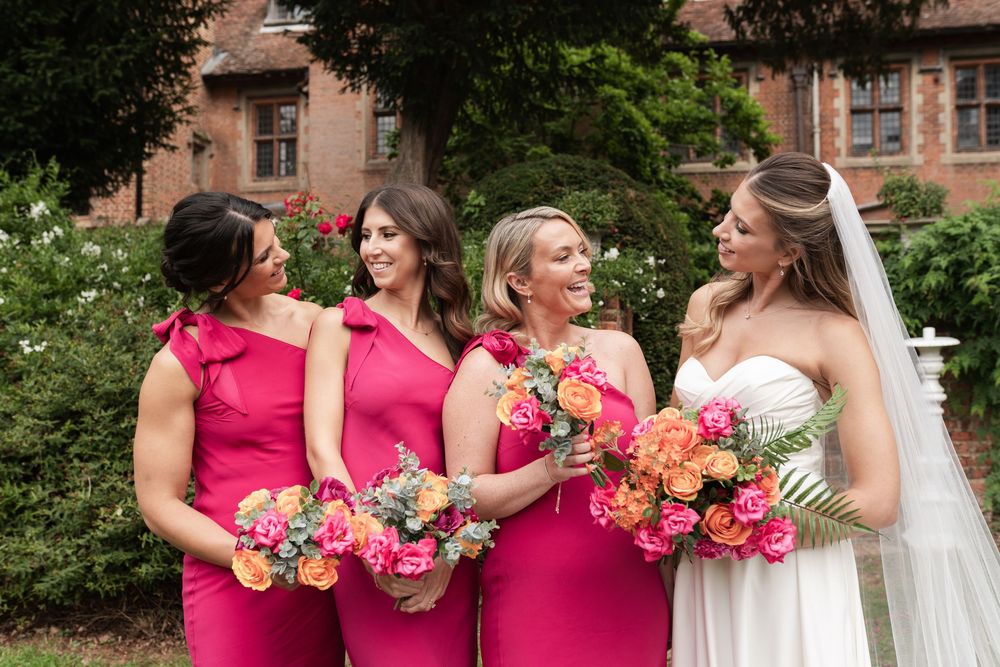 Bridesmaids in bright pink dresses gather around the bride for photos during an outdoor wedding ceremony.