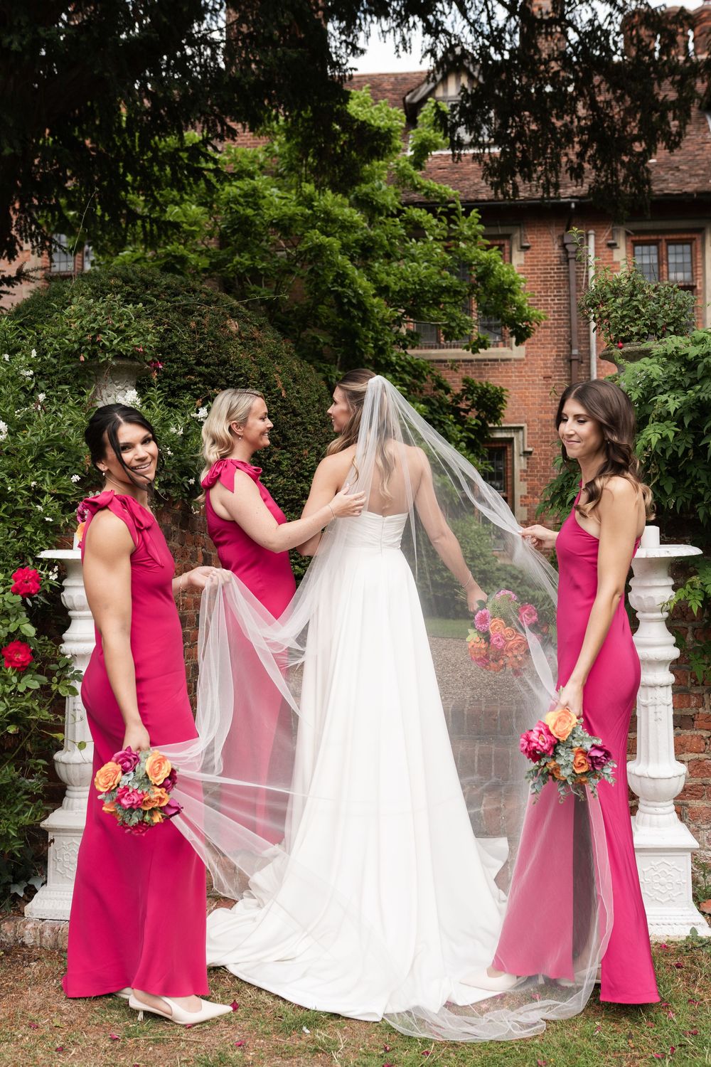 Wedding party in bright pink bridesmaid dresses gather on garden steps with bridal veil flowing in the breeze.